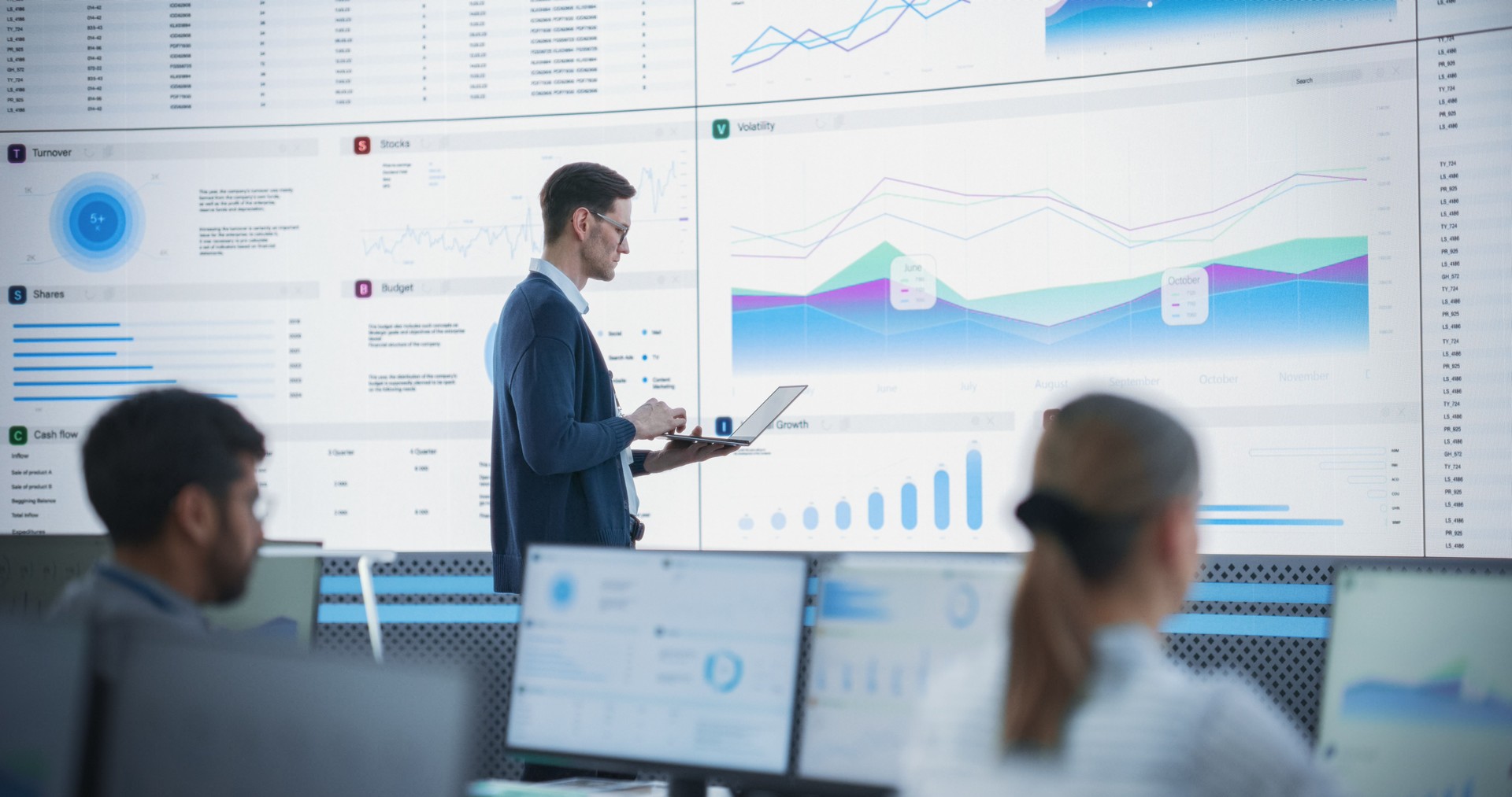 Male Senior Financial Analyst Reviewing Information And Reports on Big Digital Screen in Modern Monitoring Office. Diverse Employees Working Behind Desktop Computers In Investment Management Company.